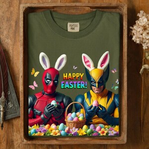 May include: Olive green t-shirt with a graphic featuring Deadpool and Wolverine wearing bunny ears and holding Easter eggs. The words "HAPPY EASTER!" are in rainbow colors. A basket of eggs and flowers are at the bottom.
