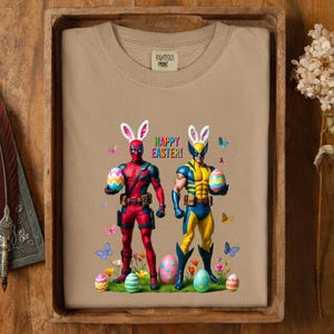 Puede incluir: Una sudadera beige con cuello redondo con un gr&aacute;fico colorido de dos superh&eacute;roes disfrazados de conejos de Pascua, sosteniendo huevos decorados. Las palabras "HAPPY EASTER!" est&aacute;n impresas encima de ellos, con mariposas y flores.
