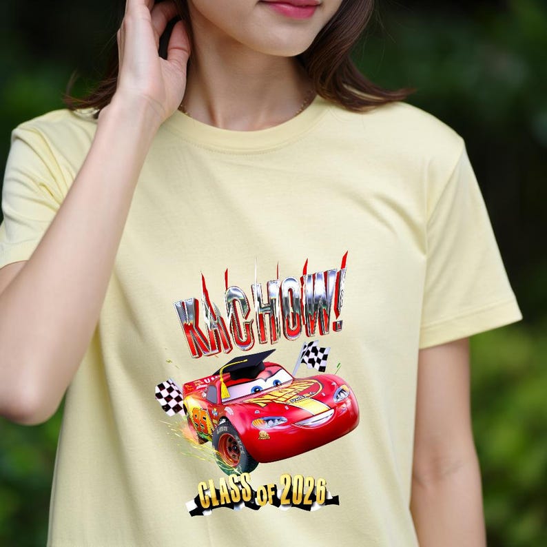 K&ouml;nnte beinhalten: Hellgelbes T-Shirt mit einer Grafik eines roten Rennwagens, dem Text "KACHOW!" und "Class of 2026". Das Auto tr&auml;gt eine Abschlusskappe und hat karierte Flaggen.