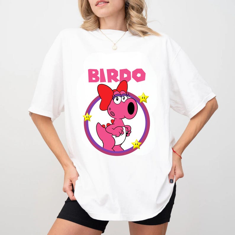 May include: A white t-shirt featuring a graphic of the pink character Birdo with a red bow, enclosed in a purple circular frame. The text BIRDO is printed in pink above the character, accompanied by three small yellow stars surrounding the circle.