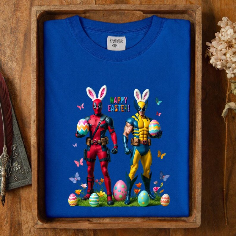Puede incluir: Sudadera azul real con Deadpool y Wolverine disfrazados de conejitos de Pascua, sosteniendo huevos decorados. Las palabras "HAPPY EASTER!" est&aacute;n impresas encima de los personajes. La sudadera est&aacute; en una bandeja de madera.