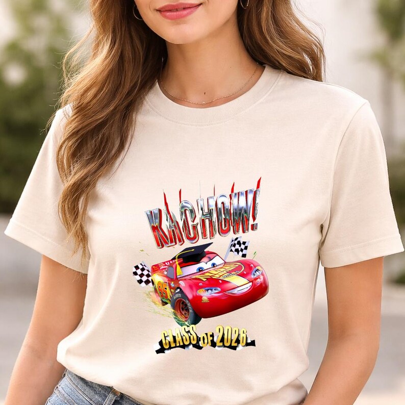 K&ouml;nnte beinhalten: Beiges T-Shirt mit einem roten Cartoon-Rennwagen, dem Text "Kachow!" und "Class of 2026." Das Auto tr&auml;gt eine Abschlusskappe und hat karierte Flaggen. Das Design ist mittig auf der Vorderseite des Shirts platziert.