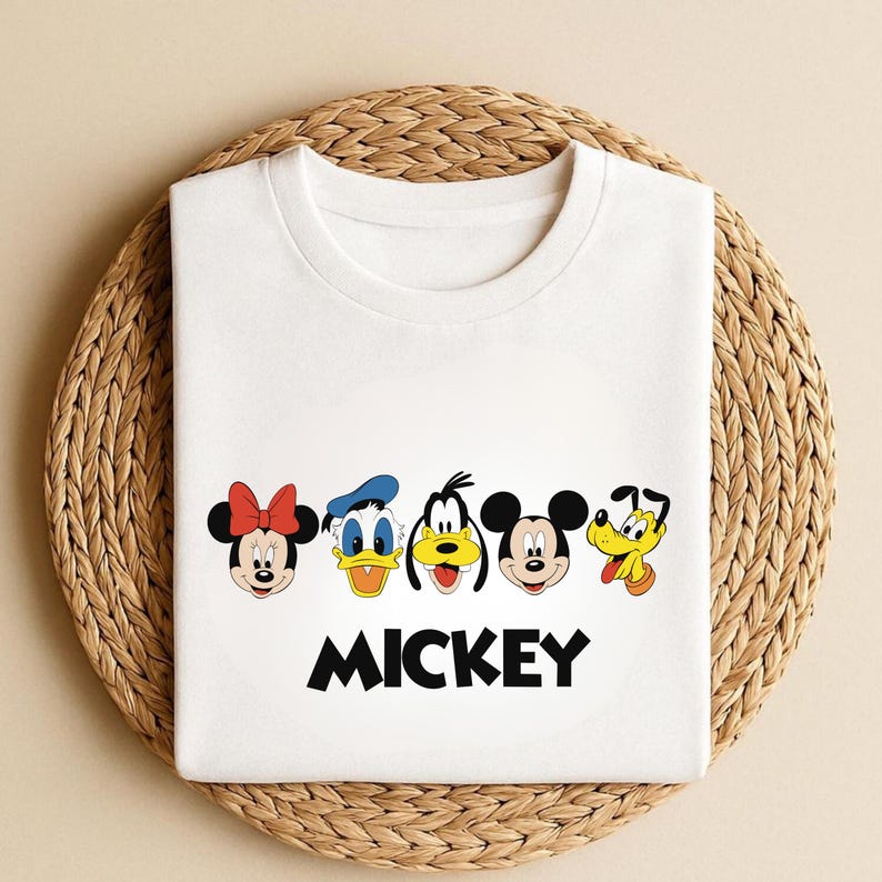 May include: White t-shirt featuring cartoon character faces of Minnie Mouse, Donald Duck, Goofy, Mickey Mouse, and Pluto. The word "MICKEY" is printed in black block letters below the characters. The shirt is folded on a woven, circular surface.