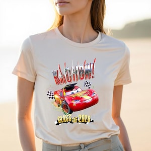 K&ouml;nnte beinhalten: Beigefarbenes T-Shirt mit einem Aufdruck eines roten Rennwagens, dem Text "Kachow!" und "Class of 2026". Der Rennwagen hat eine Abschlusskappe und Zielflaggen. Das Design ist in Rot, Gelb und Schwarz gehalten.