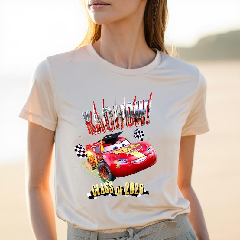 K&ouml;nnte beinhalten: Beigefarbenes T-Shirt mit einem Aufdruck eines roten Rennwagens, dem Text "Kachow!" und "Class of 2026". Der Rennwagen hat eine Abschlusskappe und Zielflaggen. Das Design ist in Rot, Gelb und Schwarz gehalten.