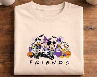May include: A beige crew neck t-shirt featuring a graphic print of cartoon characters dressed in Halloween costumes, including skeleton outfits, witch hats, and capes, surrounded by pumpkins, bats, and spiderwebs, with the text FRIENDS below.