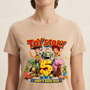 May include: A beige t-shirt featuring a colorful Toy Story graphic with Woody, Buzz Lightyear, Jessie, Rex, Slinky Dog, Hamm, Bullseye, and green army men. The design includes the text Toy Story, a large number 5, and Andy's Room 2026.