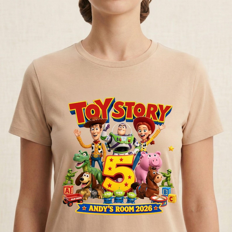 May include: A beige t-shirt featuring a colorful Toy Story graphic with Woody, Buzz Lightyear, Jessie, Rex, Slinky Dog, Hamm, Bullseye, and green army men. The design includes the text Toy Story, a large number 5, and Andy's Room 2026.