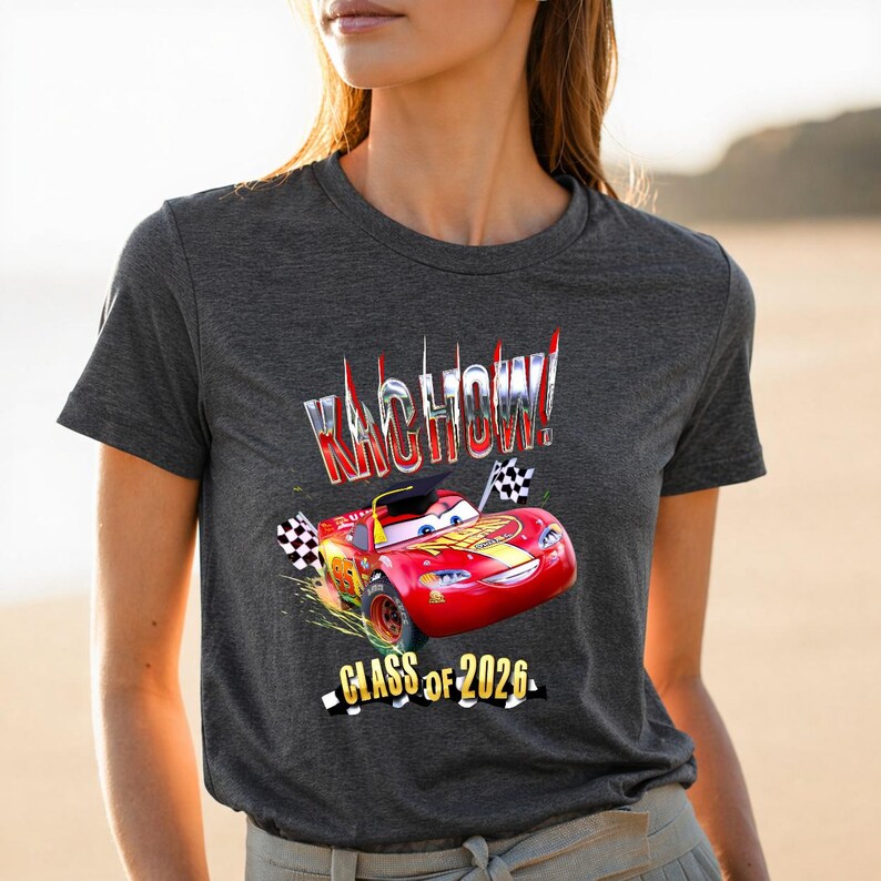 K&ouml;nnte beinhalten: Dunkelgraues T-Shirt mit einem roten Cartoon-Rennwagen, dem Text "KACHOW!" und "CLASS OF 2026." Das Auto hat eine Abschlusskappe und karierte Flaggen. Das T-Shirt hat kurze &Auml;rmel.