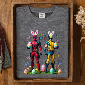 Puede incluir: Una sudadera gris con figuras de Deadpool y Wolverine con orejas de conejo y sosteniendo huevos de Pascua. Las palabras "Happy Easter!" est&aacute;n encima de ellos. Mariposas y huevos de Pascua est&aacute;n esparcidos alrededor de las figuras. La etiqueta "Righteous Print" es visible.