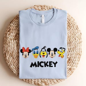 May include: A light blue t-shirt featuring cartoon character faces including Minnie Mouse, Donald Duck, Goofy, Mickey Mouse, and Pluto. The word "MICKEY" is printed in black block letters below the characters.