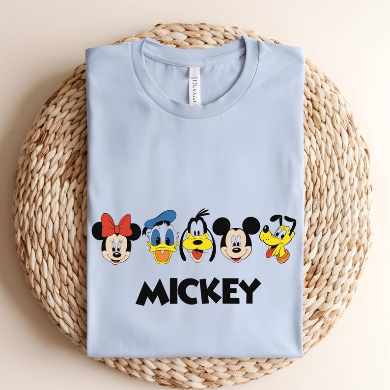 May include: A light blue t-shirt featuring cartoon character faces including Minnie Mouse, Donald Duck, Goofy, Mickey Mouse, and Pluto. The word "MICKEY" is printed in black block letters below the characters.