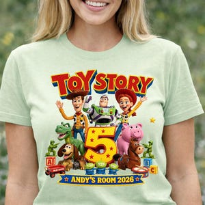 May include: A light green t-shirt featuring a colorful Toy Story graphic with characters Woody, Buzz Lightyear, Jessie, Rex, Slinky Dog, Hamm, Bullseye, and green army men surrounding a large yellow number 5 and the text Toy Story 2026.