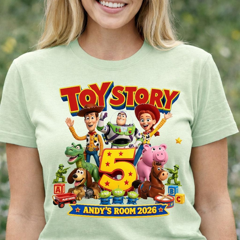 May include: A light green t-shirt featuring a colorful Toy Story graphic with characters Woody, Buzz Lightyear, Jessie, Rex, Slinky Dog, Hamm, Bullseye, and green army men surrounding a large yellow number 5 and the text Toy Story 2026.