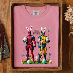 Puede incluir: Sudadera rosa con los personajes de Deadpool y Wolverine con orejas de conejo, sosteniendo huevos de Pascua. Las palabras "HAPPY EASTER!" est&aacute;n impresas encima de los personajes. La sudadera est&aacute; en una bandeja de madera.