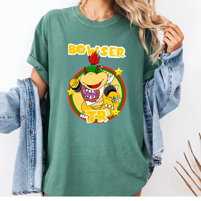 May include: A sage green short-sleeve t-shirt featuring a graphic print of the character Bowser Jr. inside a yellow and red circular emblem. The text "BOWSER JR." is printed in bold yellow letters above and below the character illustration.