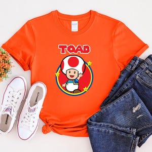 May include: A bright orange short-sleeved t-shirt featuring a graphic of the character Toad from the Super Mario series. The design includes the word TOAD in white block letters above a red circle, yellow stars, and a pair of blue denim jeans.