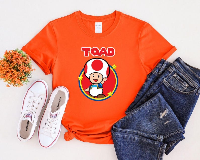 May include: A bright orange short-sleeved t-shirt featuring a graphic of the character Toad from the Super Mario series. The design includes the word TOAD in white block letters above a red circle, yellow stars, and a pair of blue denim jeans.