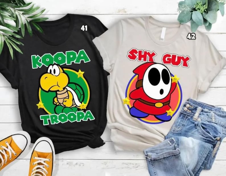 May include: A black t-shirt with a yellow Koopa Troopa character graphic and the text KOOPA TROOPA, and a light beige t-shirt with a red Shy Guy character graphic and the text SHY GUY, both featuring small yellow stars and numbered 41 and 42.
