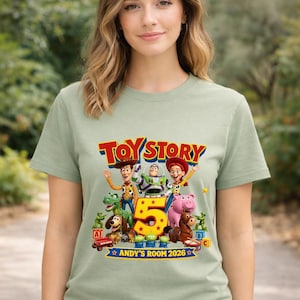 May include: A light sage green t-shirt featuring a colorful Toy Story graphic with Woody, Buzz Lightyear, Jessie, Rex, Slinky Dog, Hamm, Bullseye, and green army men surrounding a large yellow number 5, with text reading TOY STORY and ANDY'S ROOM 2026.