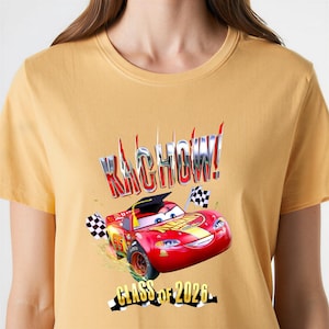 K&ouml;nnte beinhalten: Senfgelbes T-Shirt mit einer Grafik von Lightning McQueen aus dem Film Cars. Das Design beinhaltet das Wort "KACHOW!" in einer chrom&auml;hnlichen Schriftart, eine Abschlusskappe und den Text "CLASS OF 2026".
