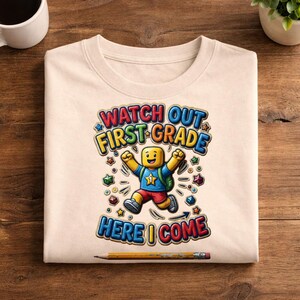 May include: A beige t-shirt featuring a graphic of a yellow block-style character wearing a blue shirt with a star and red shorts, jumping with arms raised. The text reads WATCH OUT FIRST GRADE HERE I COME, surrounded by colorful star shapes.