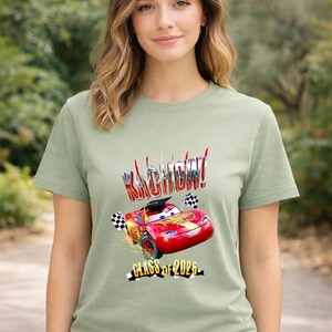 K&ouml;nnte beinhalten: Salbeigr&uuml;nes T-Shirt mit einer Grafik eines roten Rennwagens, dem Text "Kachow!" und "Class of 2026". Das Auto hat eine Abschlusskappe und Schachflaggen. Das Design ist in Rot, Gelb und Schwarz.