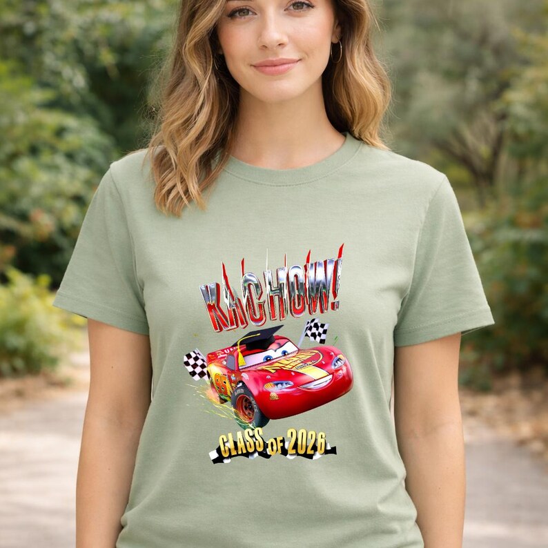 K&ouml;nnte beinhalten: Salbeigr&uuml;nes T-Shirt mit einer Grafik eines roten Rennwagens, dem Text "Kachow!" und "Class of 2026". Das Auto hat eine Abschlusskappe und Schachflaggen. Das Design ist in Rot, Gelb und Schwarz.