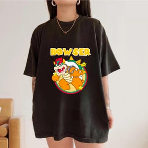 May include: A black short-sleeve t-shirt featuring a graphic of the character Bowser from the Super Mario franchise. The design includes the yellow text BOWSER above the character, who is centered within a circular frame with three yellow stars.