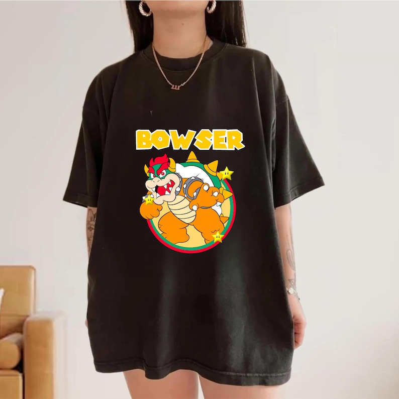 May include: A black short-sleeve t-shirt featuring a graphic of the character Bowser from the Super Mario franchise. The design includes the yellow text BOWSER above the character, who is centered within a circular frame with three yellow stars.