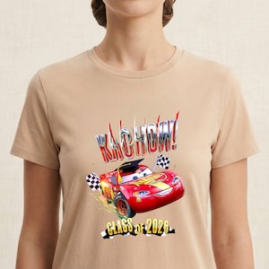 K&ouml;nnte beinhalten: Beiges T-Shirt mit einem roten Cartoon-Rennwagen, dem Text "KACHOW!" und "CLASS OF 2026". Das Auto tr&auml;gt eine Abschlusskappe und hat karierte Flaggen.