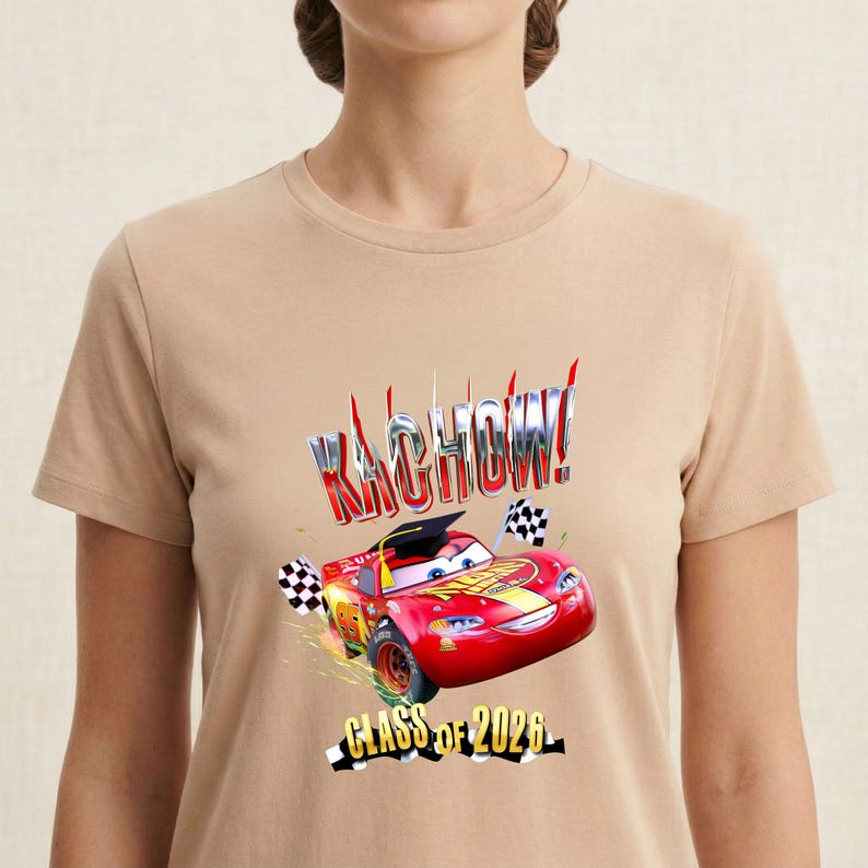 K&ouml;nnte beinhalten: Beiges T-Shirt mit einem roten Cartoon-Rennwagen, dem Text "KACHOW!" und "CLASS OF 2026". Das Auto tr&auml;gt eine Abschlusskappe und hat karierte Flaggen.