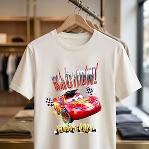 K&ouml;nnte beinhalten: Cremefarbenes T-Shirt mit einem roten Rennwagen-Motiv und dem Text "KACHOW!" in einer stilisierten Schriftart. Das Auto tr&auml;gt eine Abschlusskappe und wird von karierten Flaggen flankiert. Der Text "CLASS OF 2026" steht darunter.