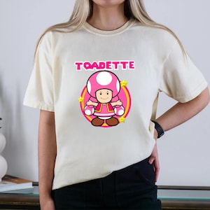 May include: A cream-colored short-sleeve t-shirt featuring a graphic of the character Toadette. The design includes the text TOADETTE in pink letters above the character, who is centered within a pink circle surrounded by small yellow stars.