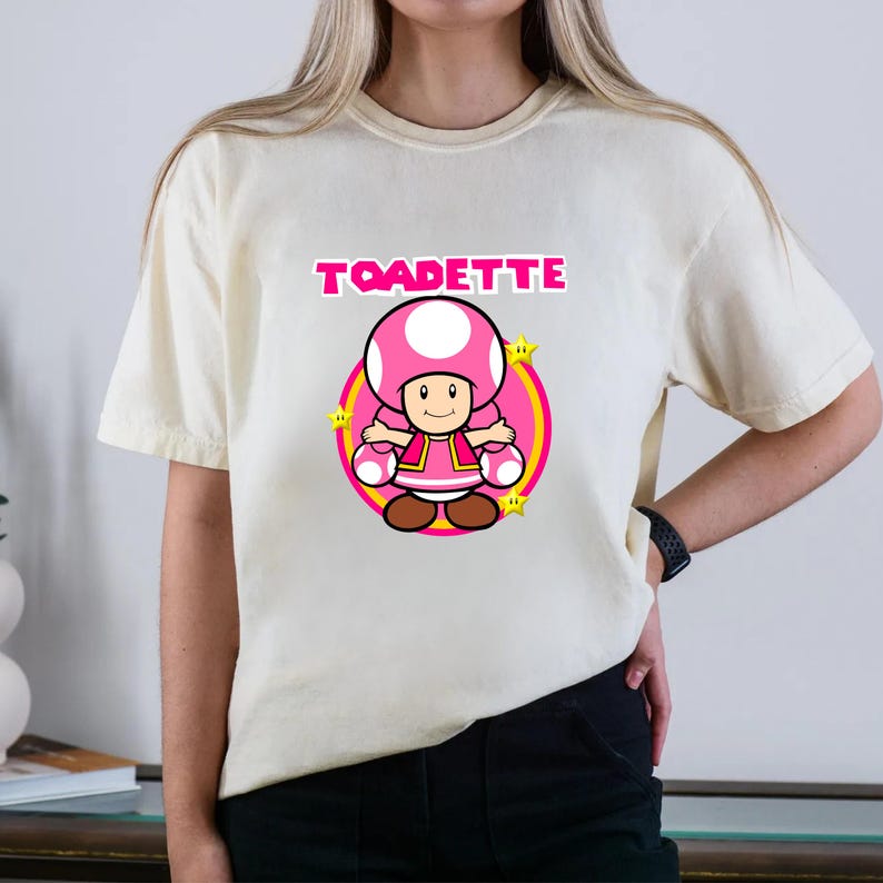 May include: A cream-colored short-sleeve t-shirt featuring a graphic of the character Toadette. The design includes the text TOADETTE in pink letters above the character, who is centered within a pink circle surrounded by small yellow stars.