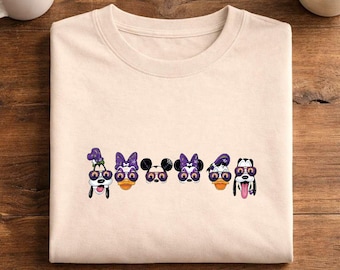 May include: A light beige crew neck t-shirt featuring a graphic print of six cartoon character heads arranged in a horizontal row. Each character wears purple-tinted sunglasses with a small pumpkin icon on the lenses, set against a neutral background.