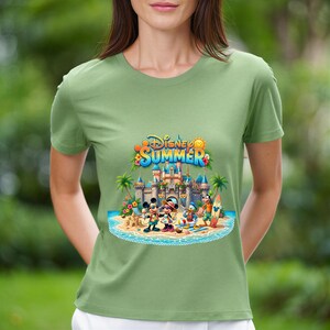May include: A sage green short-sleeve t-shirt featuring a colorful graphic of Disney characters at a beach with a castle in the background. The text Disney SUMMER is printed in blue and yellow above the castle, with palm trees and sand.