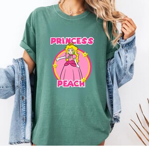 May include: A sage green short-sleeve t-shirt featuring a graphic of Princess Peach in a pink dress inside a yellow-bordered pink circle. The text PRINCESS PEACH is printed in bold pink letters above and below the character, with yellow stars.