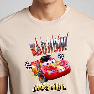K&ouml;nnte beinhalten: Beiges T-Shirt mit einer Grafik von Lightning McQueen aus dem Film Cars, der eine Abschlusskappe tr&auml;gt. Die Texte "KACHOW!" und "CLASS OF 2026" sind ebenfalls abgebildet. Das Design umfasst karierte Flaggen und Renndetails.