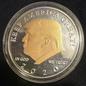 以下が含まれることがあります： 「KEEP AMERICA GREAT!」と「IN GOD WE TRUST」の文字が入った銀と金のコイン。人物の横顔と2020年の文字が描かれています。