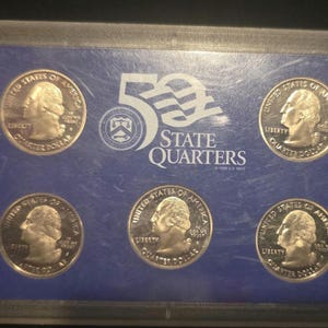 May include: A set of five United States quarter coins in a clear plastic case. The coins feature a profile of George Washington and the words "Liberty" and "Quarter Dollar." The case has a blue background with the text "50 State Quarters."