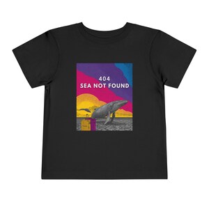 以下が含まれることがあります： クジラのグラフィックと「404 SEA NOT FOUND」のテキストがプリントされた黒い幼児用Tシャツ。グラフィックにはクジラ、2つのスーツケース、紫、青、黄、オレンジの色合いのカラフルな背景が含まれています。