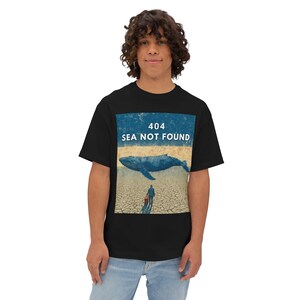 May include: Black t-shirt featuring a graphic with a whale on cracked earth, a person with a suitcase, and the text "404 SEA NOT FOUND". The design is in shades of blue, beige, and brown.