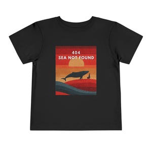 以下が含まれることがあります： 黒い幼児用Tシャツ。クジラ、人物、そして「404 SEA NOT FOUND」のテキストが夕焼けを背景に描かれたグラフィックが特徴です。デザインはオレンジ、茶色、青の色合いを使用しています。