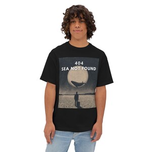 May include: Black t-shirt featuring a surreal graphic with a whale, a person, and a full moon. The text "404 SEA NOT FOUND" is printed above the image. The shirt is worn by a person.