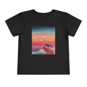 以下が含まれることがあります： 黒い幼児用Tシャツ。グラフィックは、波に乗るクジラ、人、そして「404 SEA NOT FOUND」のテキストを、カラフルなストライプの夕焼けの背景に描いています。柔らかい素材で作られたシャツです。