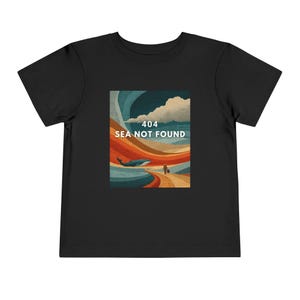 以下が含まれることがあります： クジラ、人、そして「404 SEA NOT FOUND」のテキストが特徴のグラフィックプリントが施された黒い幼児用Tシャツ。アートワークは、青いクジラと小道を歩く人を特徴とするレトロなスタイルです。