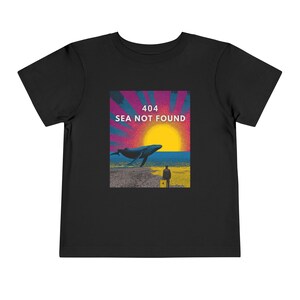 以下が含まれることがあります： クジラ、夕日、そして「404 SEA NOT FOUND」のテキストが描かれた黒い幼児用Tシャツ。デザインには、ビーチにスーツケースを持った人が含まれています。シャツは柔らかい素材で作られています。
