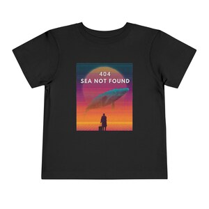 以下が含まれることがあります： 黒い幼児用Tシャツ。クジラ、スーツケースを持った人物、そして「404 SEA NOT FOUND」のテキストが描かれたグラフィックデザイン。オレンジ、紫、青の色合いを使ったレトロな夕焼けの配色です。