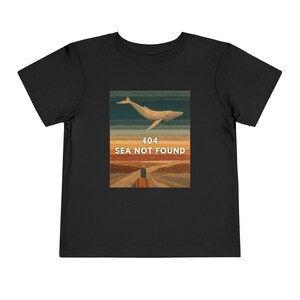 以下が含まれることがあります： 黒い幼児用Tシャツ。グラフィックデザインは、テキスト「404 SEA NOT FOUND」の上にクジラのイラストが描かれています。画像には、夕焼けを背景に、道路を歩く荷物を持った人が描かれています。